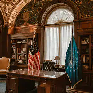 Office with American and blue flags, wooden bookshelves, and elegant decor.
